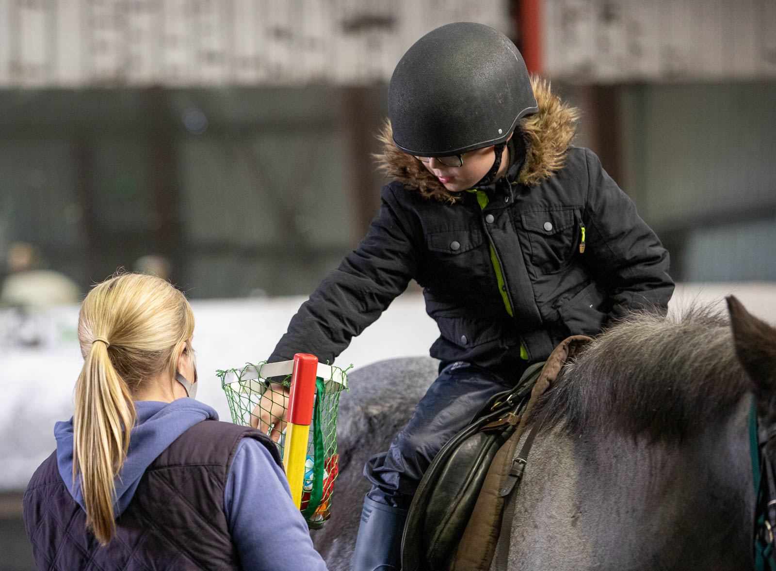 Image of young rider on a pony reaching for a toy supported by a volunteer